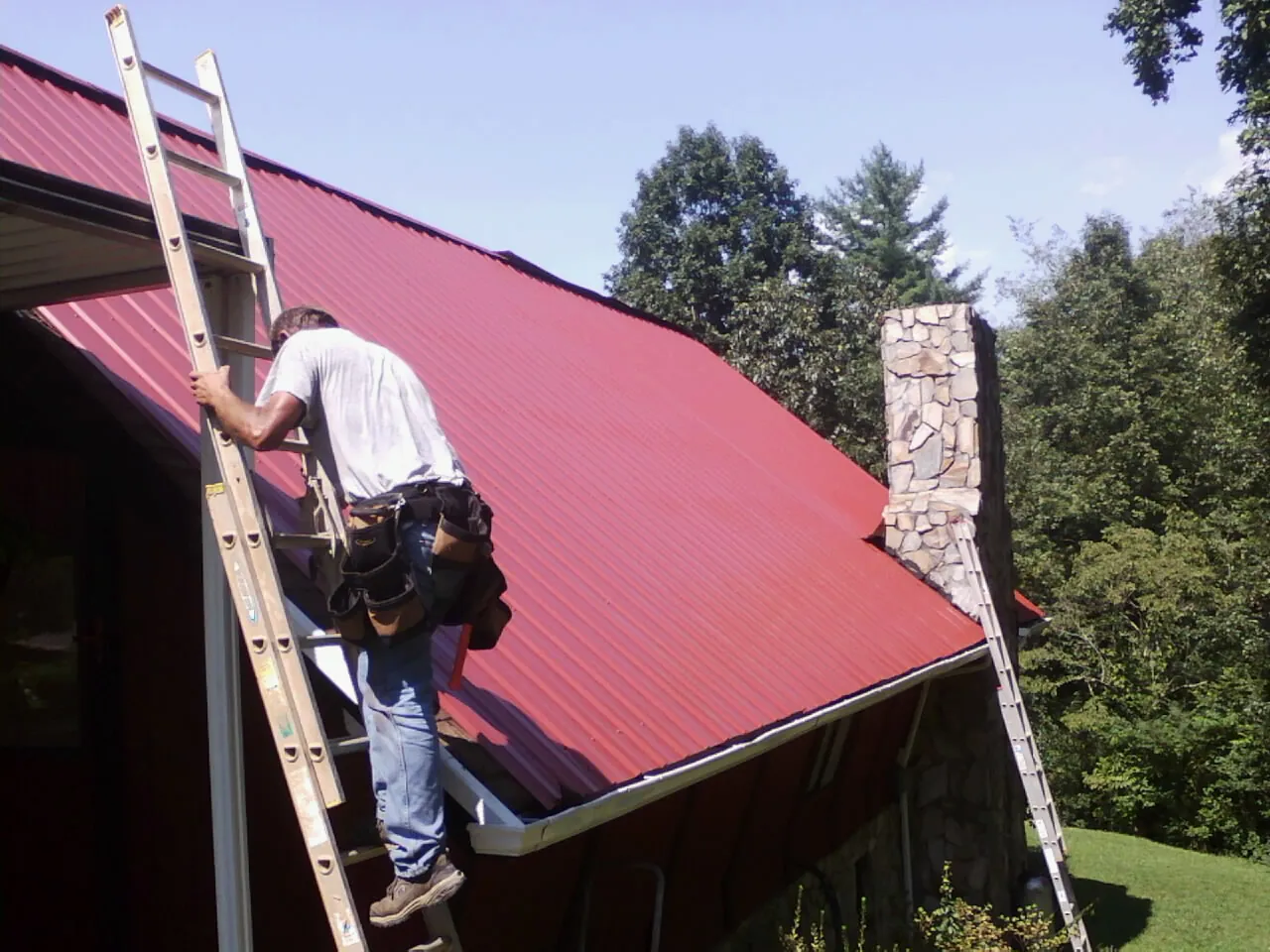 Roofer on a ladder beside a red metal roof and stone chimney — Soffit & Fascia Repair by Roofing Brothers in Bowman